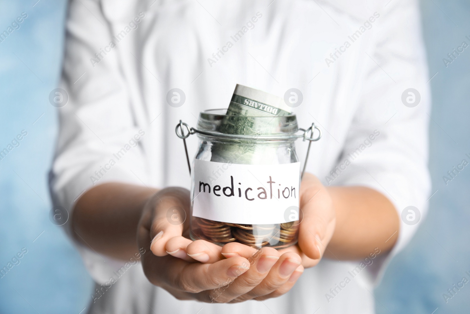 Photo of Woman holding glass jar with money and tag MEDICATION on light blue background, closeup