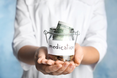 Woman holding glass jar with money and tag MEDICATION on light blue background, closeup