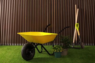 Wheelbarrow, gardening tools and plants on green grass near wood slat wall