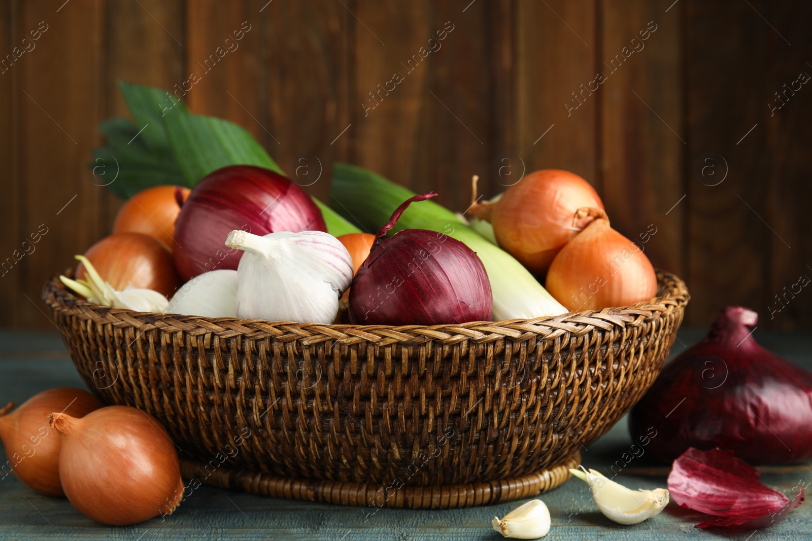 Photo of Wicker basket with fresh onion bulbs, leeks and garlic on light blue wooden table