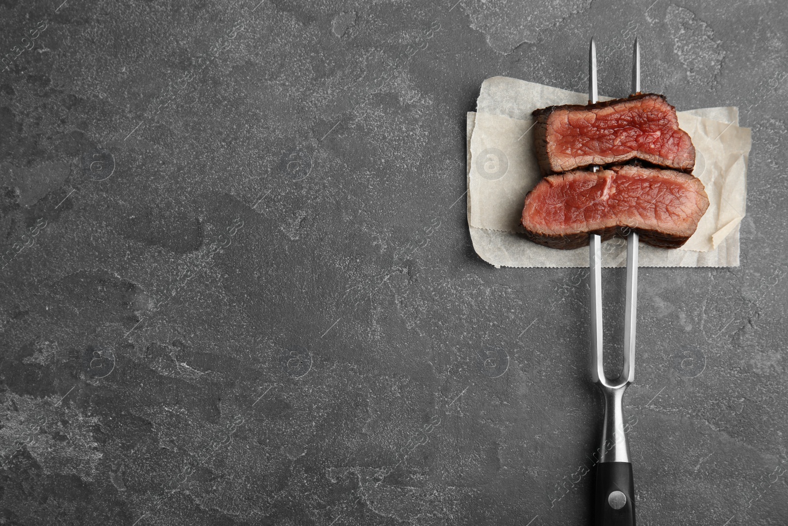 Photo of Carving fork with slices of delicious beef steak on grey table, top view. Space for text