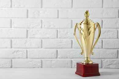 Golden trophy cup on table near white brick wall. Space for text