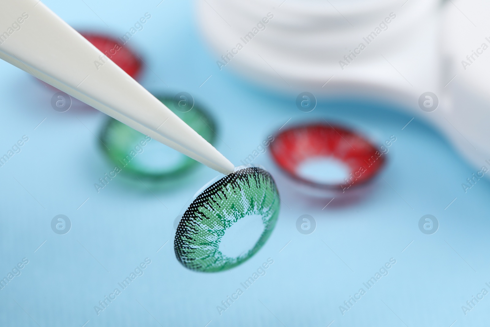 Photo of Tweezers with green contact lens on light blue background, closeup. Space for text