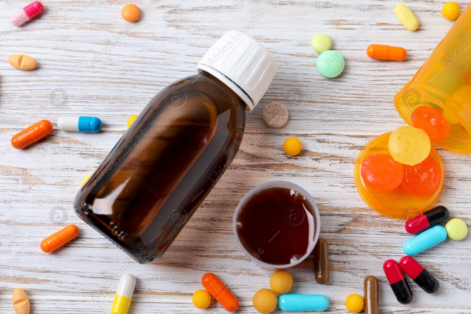 Photo of Bottle of syrup, measuring cup, lozenges and pills on white wooden table, flat lay. Cold medicine