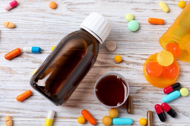 Photo of Bottle of syrup, measuring cup, lozenges and pills on white wooden table, flat lay. Cold medicine