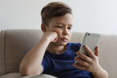 Photo of Little child with smartphone on sofa in room. Danger of internet