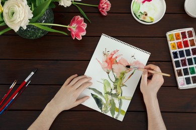 Photo of Woman painting flowers with watercolor at wooden table, top view. Creative artwork