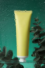 Tube with moisturizing cream and eucalyptus branches on green background, view through wet glass