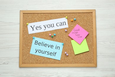Corkboard with pinned motivational quotes on white wooden background, top view