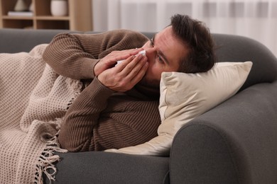 Photo of Sick man with tissue blowing nose on sofa at home. Cold symptoms