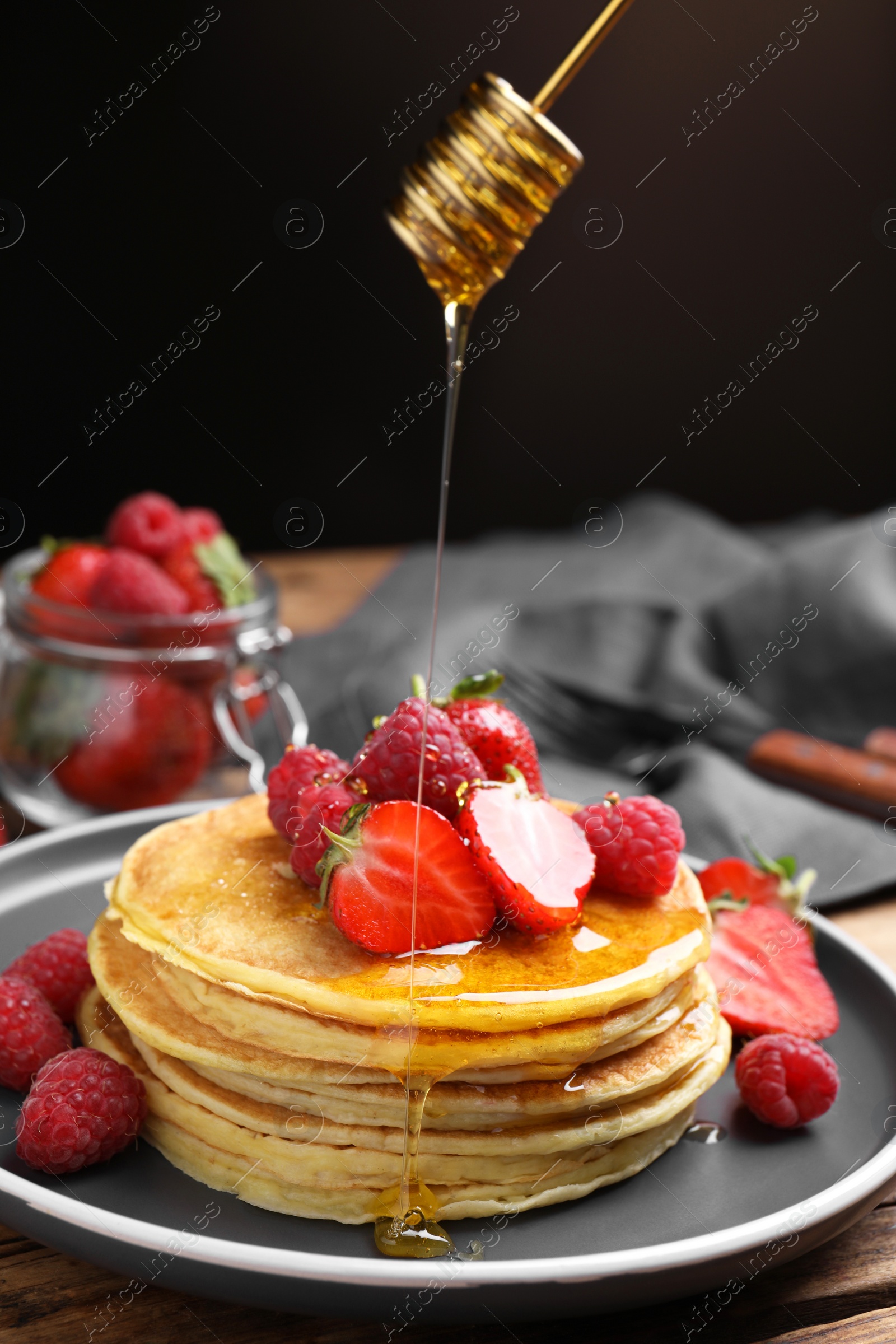 Photo of Pouring honey onto tasty pancakes with fresh berries on wooden table, closeup