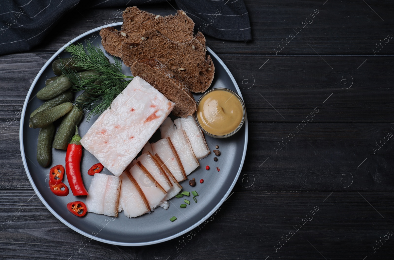 Photo of pork fatback with spices, bread and pickled cucumbers on black wooden table, flat lay. Space for text