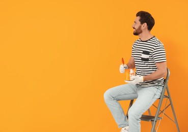 Photo of Happy designer with painting equipment sitting on folding ladder near freshly painted orange wall, space for text