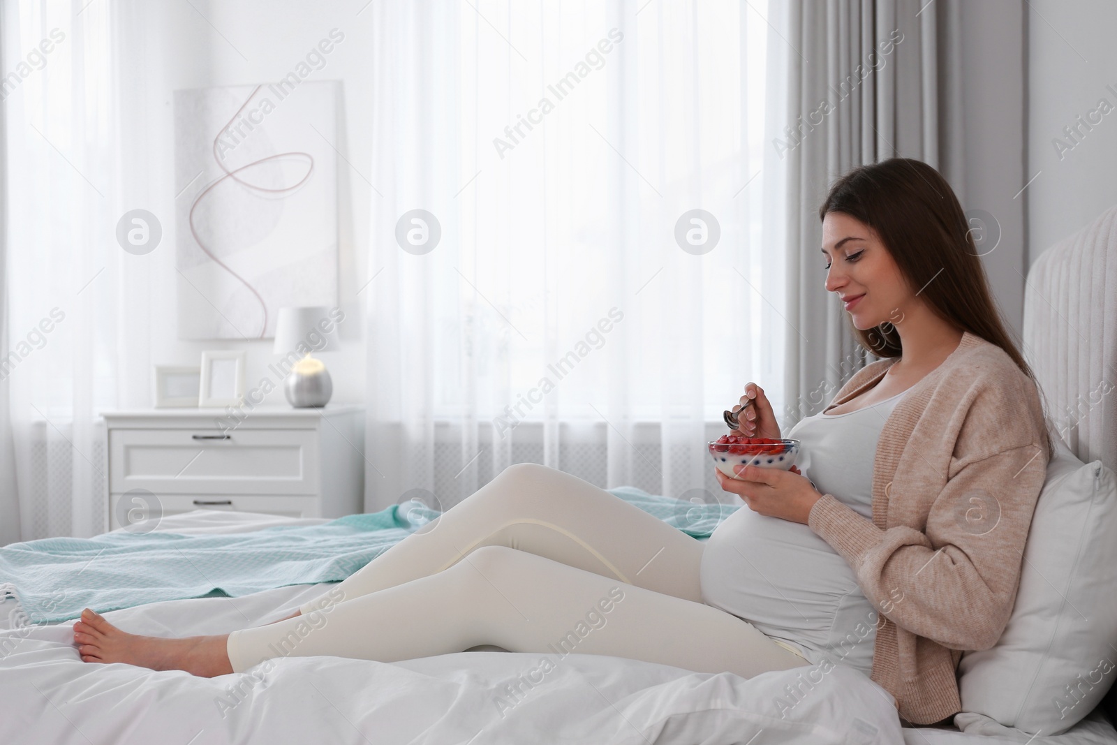 Photo of Pregnant woman eating yogurt with berries on bed at home. Healthy diet