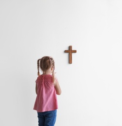 Little girl praying near light wall with cross