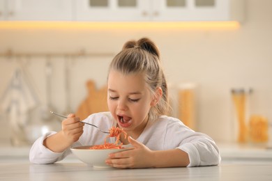 Photo of Cute little girl eating tasty pasta at table in kitchen