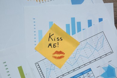 Photo of Sticky note with phrase Kiss Me and lipstick mark attached to documents, top view