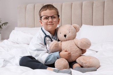 Cute little boy in pediatrician's uniform playing with stethoscope and toy bear at home