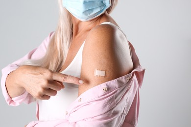 Mature woman in protective mask pointing at arm with bandage after vaccination on light background, closeup