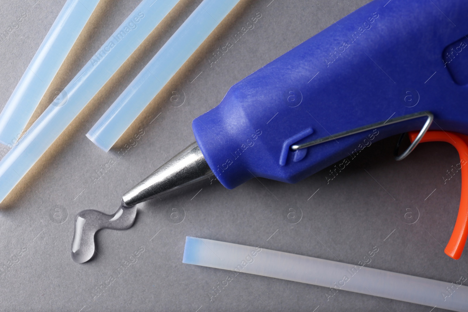 Photo of Melted glue dripping out of hot gun nozzle near sticks on grey background, closeup