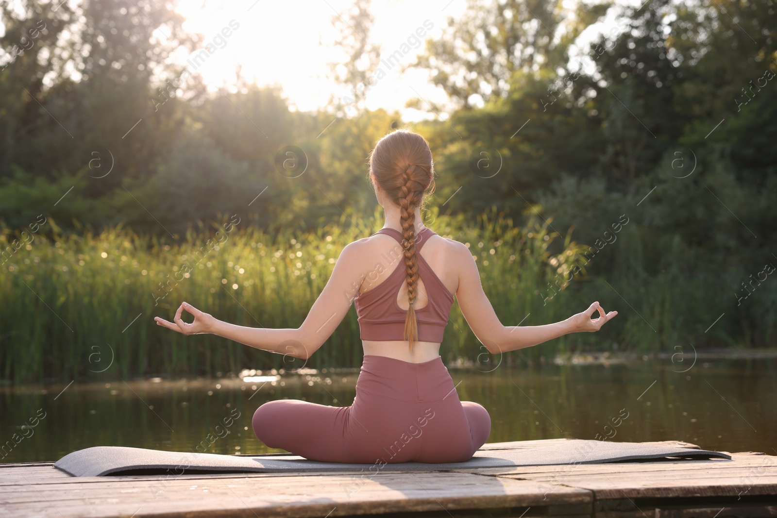 Photo of Woman practicing Padmasana on yoga mat on wooden pier near pond, back view. Lotus pose