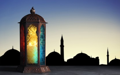 Image of Decorative Arabic lantern on stone surface and silhouette of mosque at sunset on background, space for text