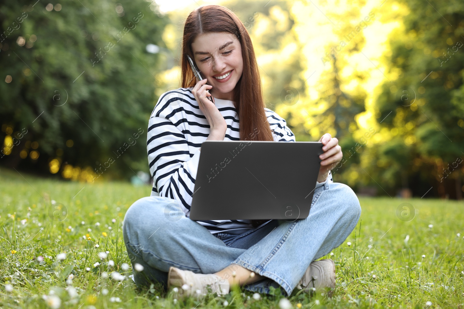 Photo of Smiling freelancer with laptop talking by smartphone on green grass outdoors. Remote job