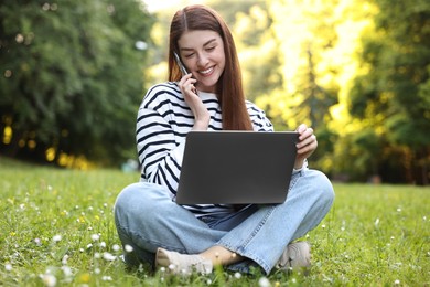 Photo of Smiling freelancer with laptop talking by smartphone on green grass outdoors. Remote job