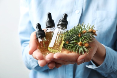 Woman holding bottles of essential oils and fir branches on white background, closeup