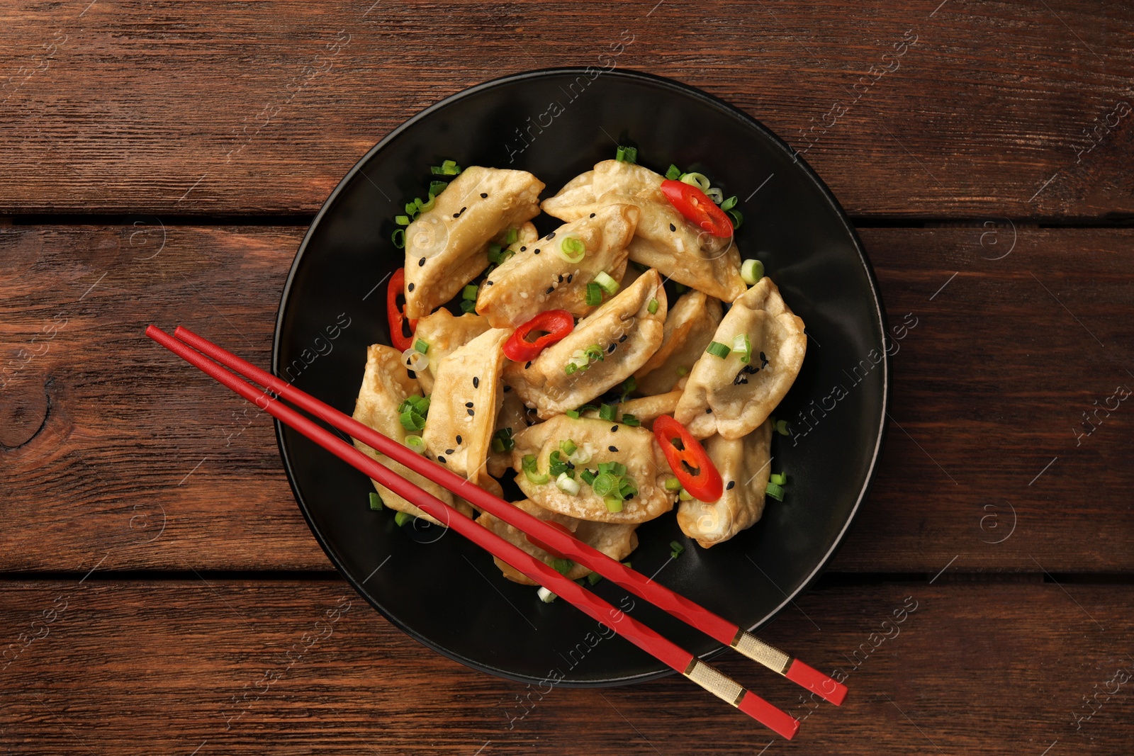 Photo of Delicious gyoza (asian dumplings) with sesame seeds, green onions, chili peppers and chopsticks on wooden table, top view