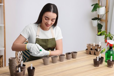 Photo of Young woman spraying water onto vegetable seeds in peat pots at wooden table indoors