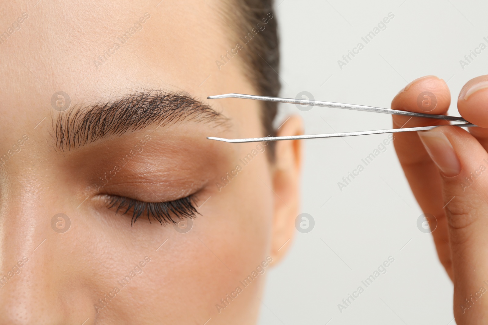 Photo of Eyebrow correction. Young woman with tweezers on light grey background, closeup