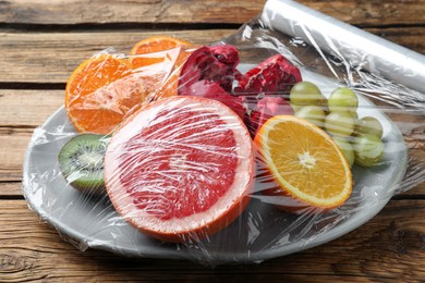 Photo of Plate of fresh fruits with plastic food wrap on wooden table, closeup