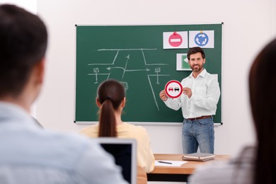 Teacher showing No Overtaking road sign to audience near chalkboard in driving school