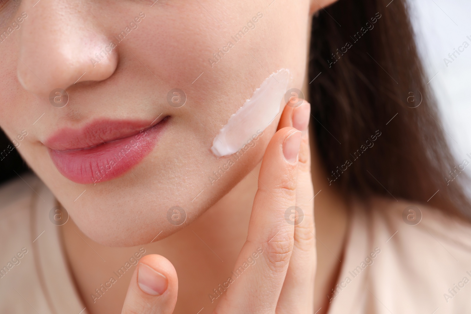 Photo of Young woman with dry skin applying cream onto her face on blurred background, closeup