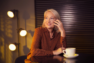 Beautiful mature woman with cup of coffee talking on smartphone indoors