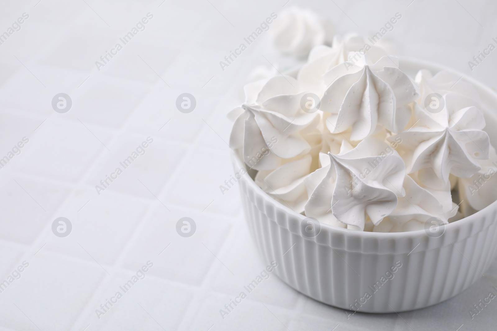 Photo of Delicious meringue cookies in bowl on white tiled table, closeup. Space for text