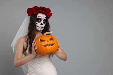 Photo of Young woman in scary bride costume with sugar skull makeup, flower crown and carved pumpkin on light grey background, space for text. Halloween celebration