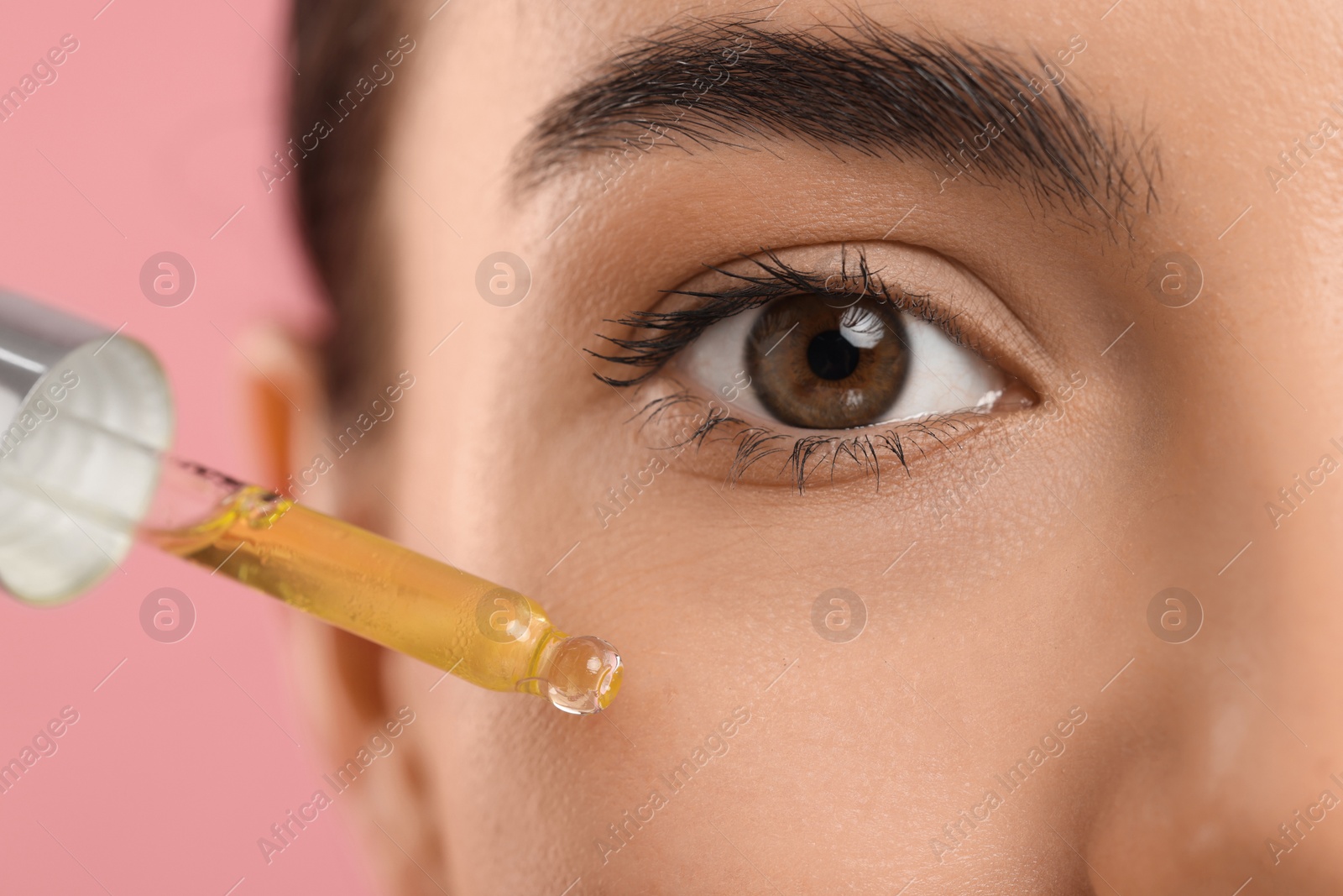 Photo of Young woman applying serum onto her face on pink background, closeup