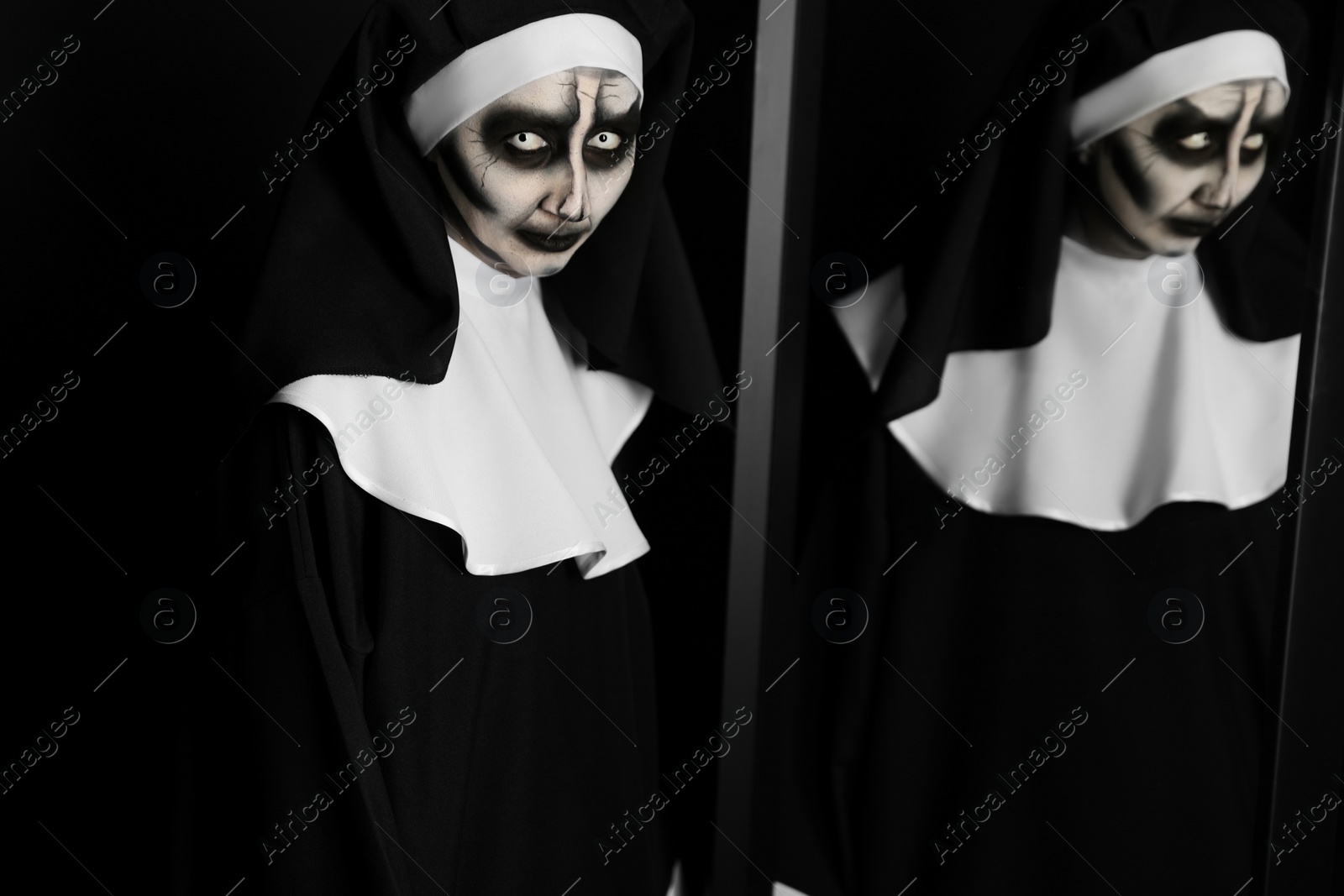 Photo of Scary devilish nun near mirror on black background. Halloween party look