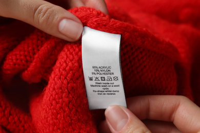 Photo of Woman reading clothing label with care symbols and material content on red knitted sweater, closeup