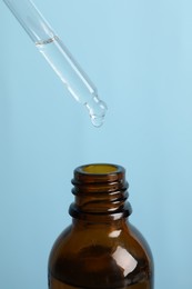 Photo of Dripping cosmetic serum from pipette into bottle on light blue background