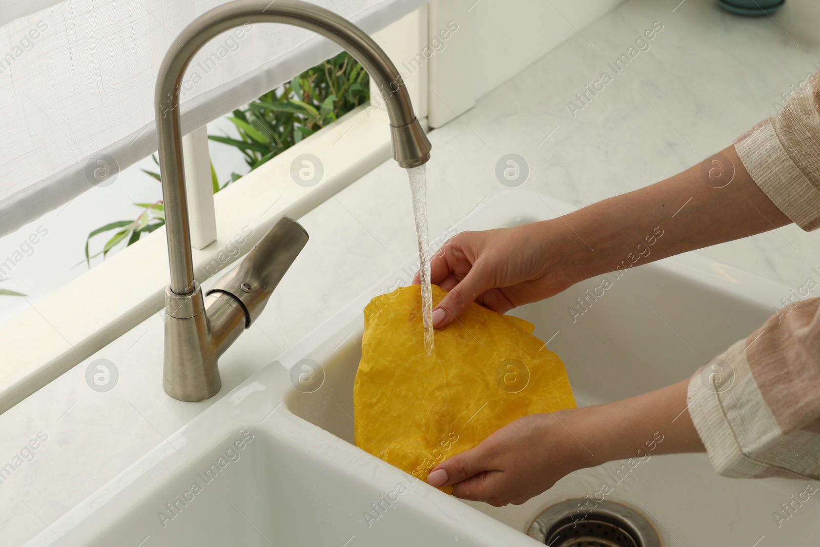 Photo of Woman washing beeswax food wrap under tap water in kitchen sink, closeup