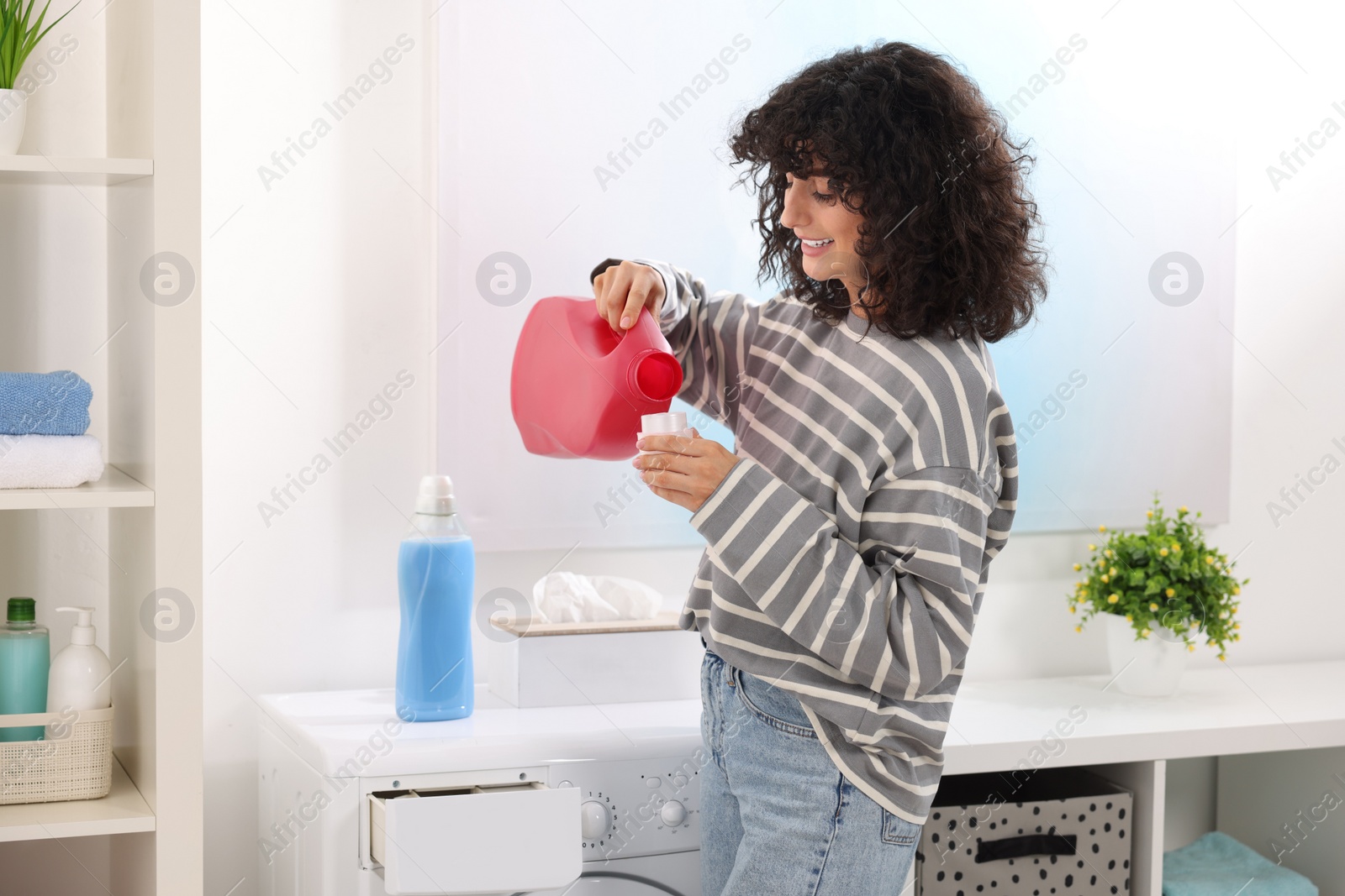 Photo of Happy woman pouring laundry detergent into cap near washing machine indoors