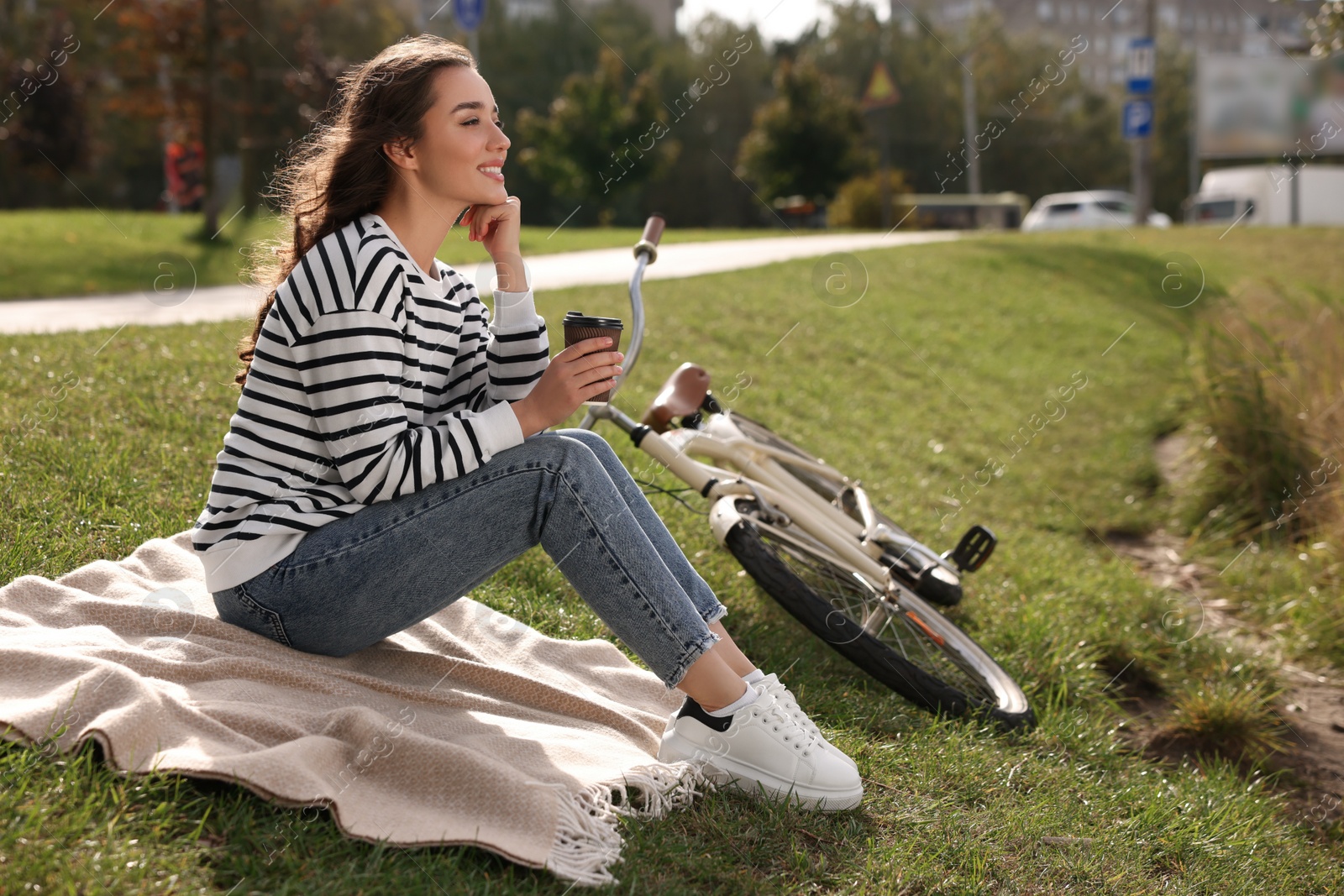 Photo of Young woman sitting on green grass and holding cup of coffee near bicycle in park, space for text