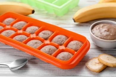 Banana puree in ice cube tray with ingredients on white wooden table, closeup