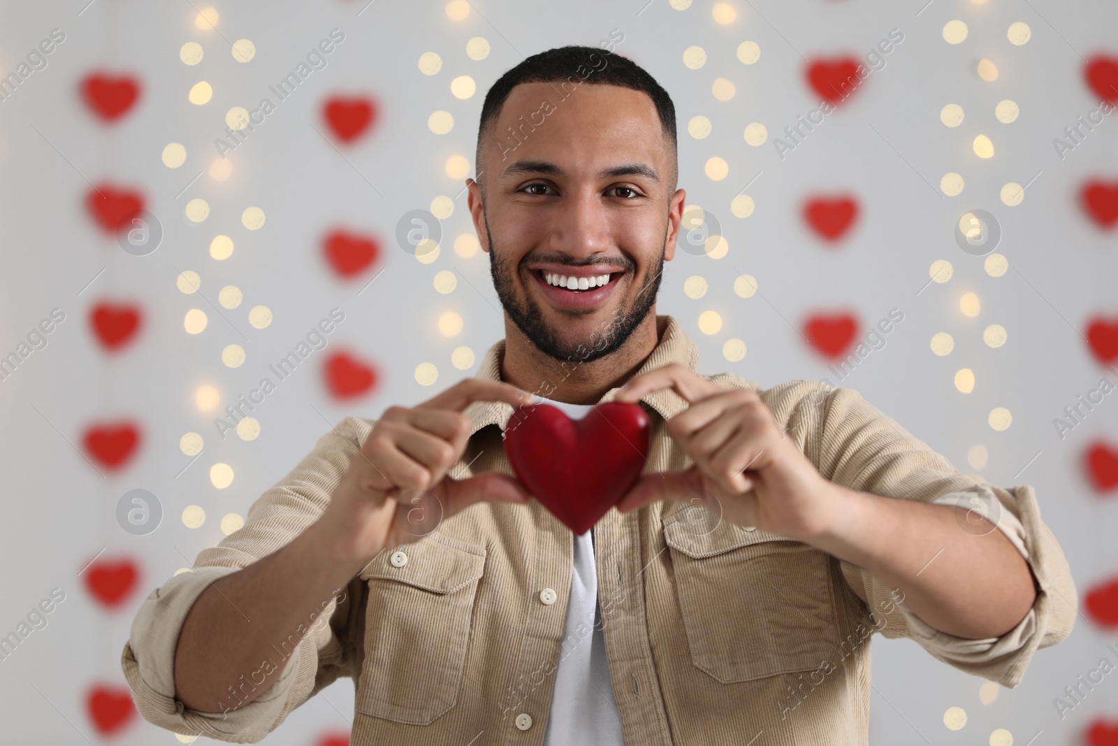 Photo of Handsome young man with red wooden heart indoors, view from camera. Valentine's day celebration in long distance relationship