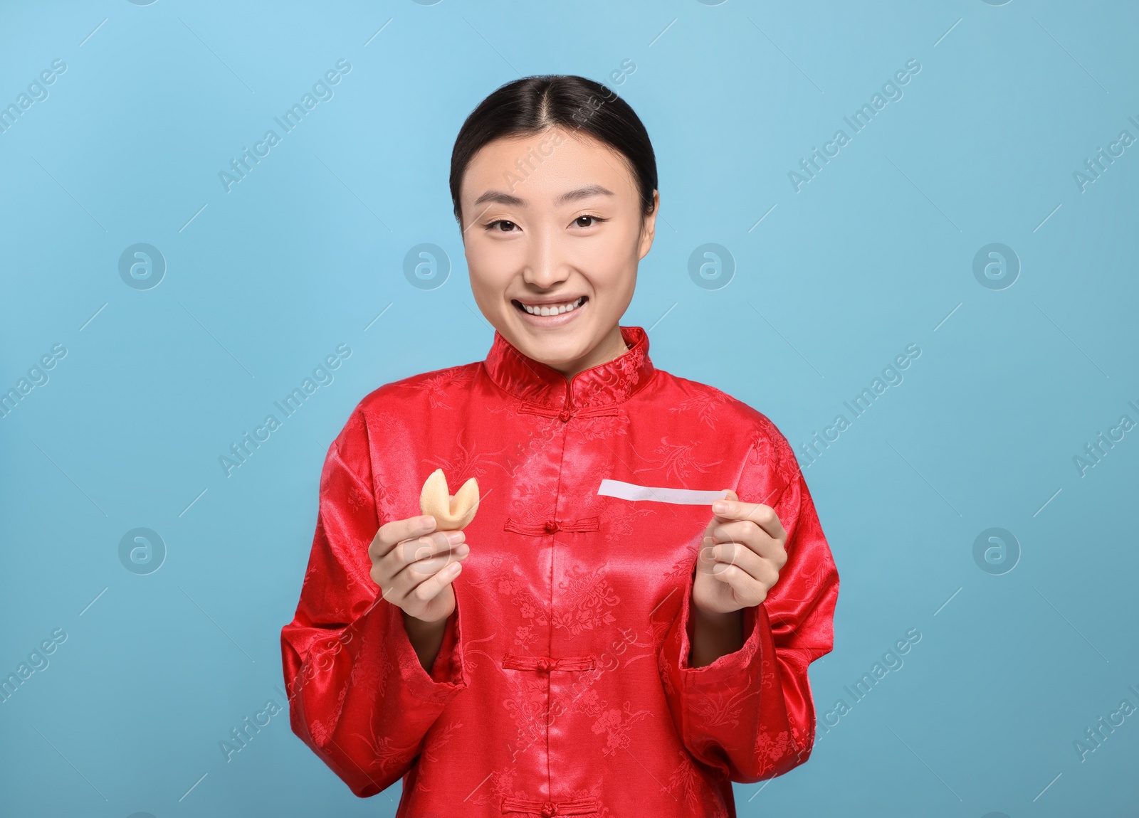 Photo of Asian woman holding tasty fortune cookie with prediction on light blue background, space for text