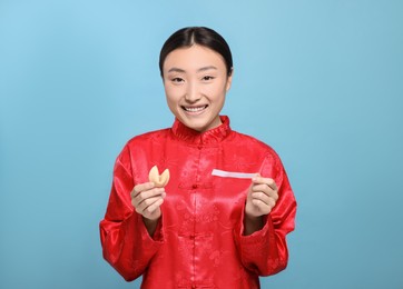 Asian woman holding tasty fortune cookie with prediction on light blue background, space for text