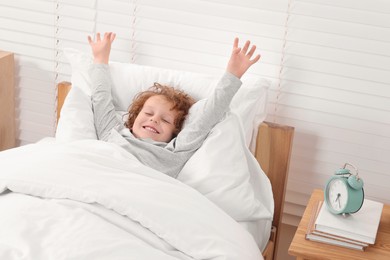 Photo of Cute little boy stretching in cosy bed near alarm clock on bedside table indoors